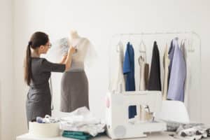 Sarta vestita di grigio con lunghi capelli che lavora su un manichino davanti c'è una scrivania con macchina da cucire e stoffe bianchi e sullo sfondo uno stand con vestiti appesi