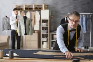 sarto con camicia celeste, gilet blu con metro al collo lavora al tavolo sullo sfondo un uomo che prova una giacca stand con vestiti appesi