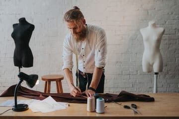 sarto con camicia bianca jeans barba e capelli raccolti che lavora ad un abito su una scrivania marrone chiara su cui c'i sono fili e una lampada, sullo sfondo due manichini mezzobusto uno bianco e uno nero