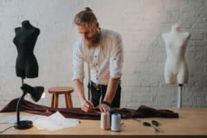 sarto con camicia bianca jeans barba e capelli raccolti che lavora ad un abito su una scrivania marrone chiara su cui c'i sono fili e una lampada, sullo sfondo due manichini mezzobusto uno bianco e uno nero